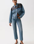 A model wearing a cropped denim jacket with a color-blocked design in medium and dark blue, paired with matching blue jeans and black pointed-toe heels, against a plain white studio background.