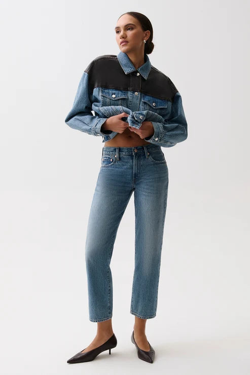 A model wearing a cropped denim jacket with a color-blocked design in medium and dark blue, paired with blue jeans and black pointed-toe heels, against a plain light gray studio background.