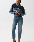 A model wearing a cropped denim jacket with a color-blocked design in medium and dark blue, paired with blue jeans and black pointed-toe heels, against a plain light gray studio background.