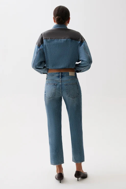 A model shown from the back wearing a cropped denim jacket with dark gray shoulder panels and blue denim body, paired with blue jeans and black heels, against a plain white studio background.