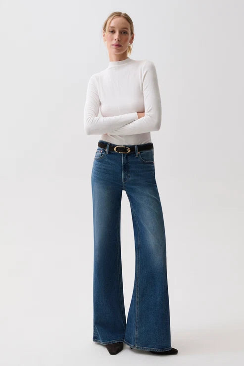 A woman modeling a white long-sleeve mock neck top and blue wide-leg jeans with a black belt, standing against a plain light gray studio background.