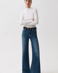 A woman modeling a white long-sleeve mock neck top and blue wide-leg jeans with a black belt, standing against a plain light gray studio background.