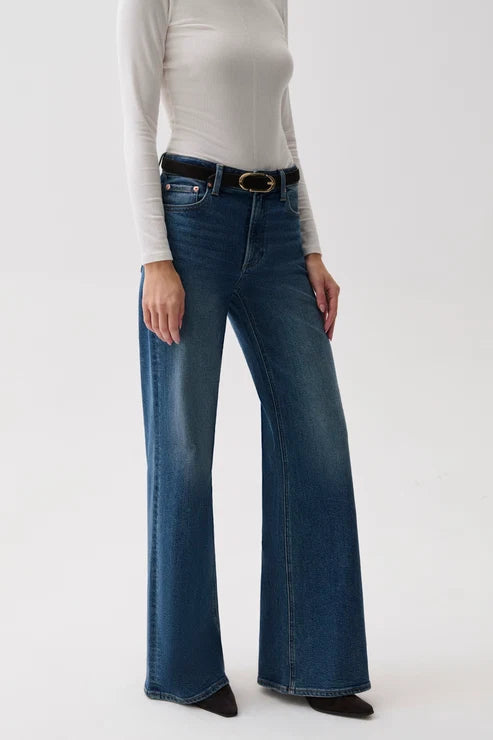 A model wearing a pair of dark blue wide-leg jeans with a black belt, paired with a white long-sleeve top and black pointed shoes, against a plain white studio background.