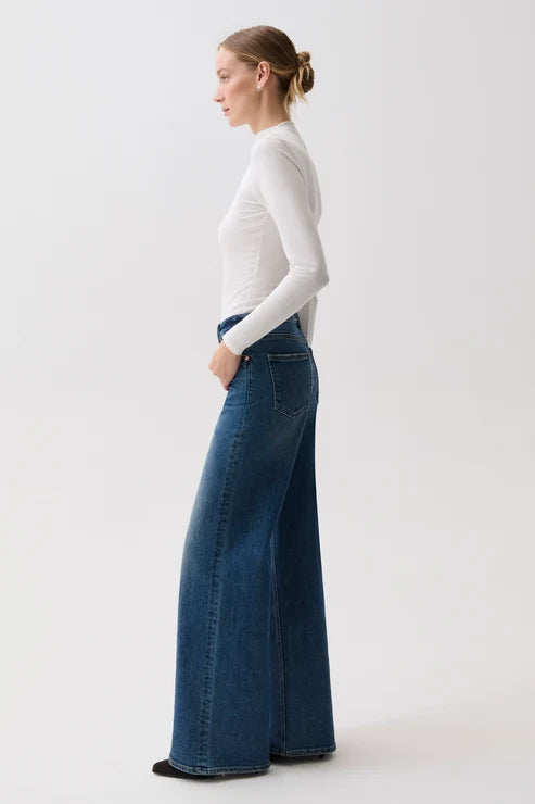 A woman in profile wearing a fitted white long-sleeve top and high-waisted, wide-leg blue denim jeans against a plain light gray studio background.