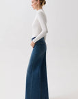 A woman in profile wearing a fitted white long-sleeve top and high-waisted, wide-leg blue denim jeans against a plain light gray studio background.