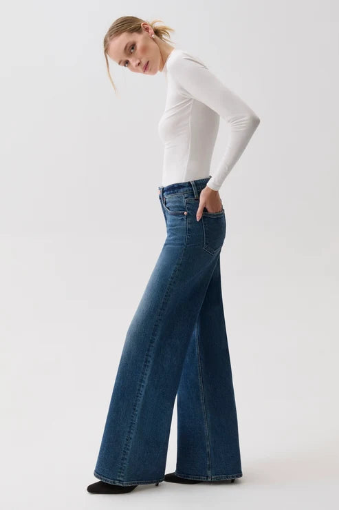 A woman in profile wearing a fitted white long-sleeve top and high-waisted, wide-leg blue denim jeans against a plain light gray studio background.