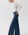 A woman in profile wearing a fitted white long-sleeve top and high-waisted, wide-leg blue denim jeans against a plain light gray studio background.