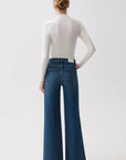 A model shown from the back wearing a white long-sleeve mock-neck top and high-waisted, wide-leg blue denim jeans against a plain white studio background.