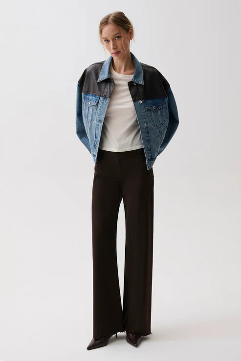 A model stands against a light gray studio background wearing a cropped denim jacket with black shoulder panels, a white ribbed top, dark wide-leg trousers, and pointed black shoes.