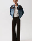 A model stands against a light gray studio background wearing a cropped denim jacket with black shoulder panels, a white ribbed top, dark wide-leg trousers, and pointed black shoes.