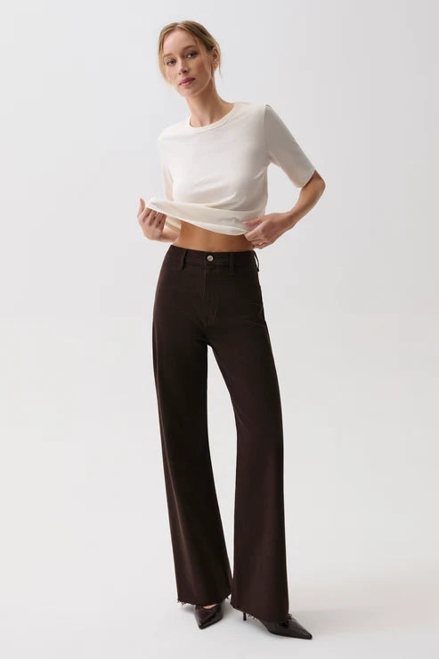 A model wearing a white short-sleeve crop top and dark brown wide-leg trousers, standing against a light gray studio background.
