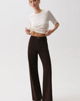 A model wearing a white short-sleeve crop top and dark brown wide-leg trousers, standing against a light gray studio background.