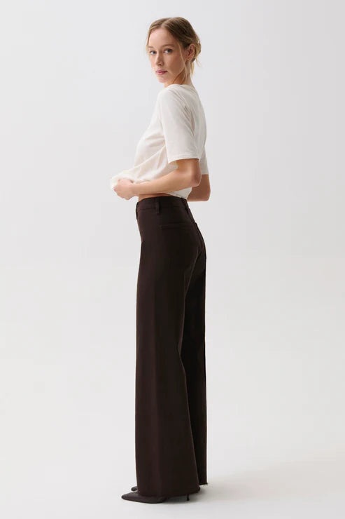 A woman stands in profile wearing a fitted white short-sleeve top and dark brown wide-leg trousers against a clean light gray studio background.