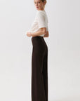 A woman stands in profile wearing a fitted white short-sleeve top and dark brown wide-leg trousers against a clean light gray studio background.