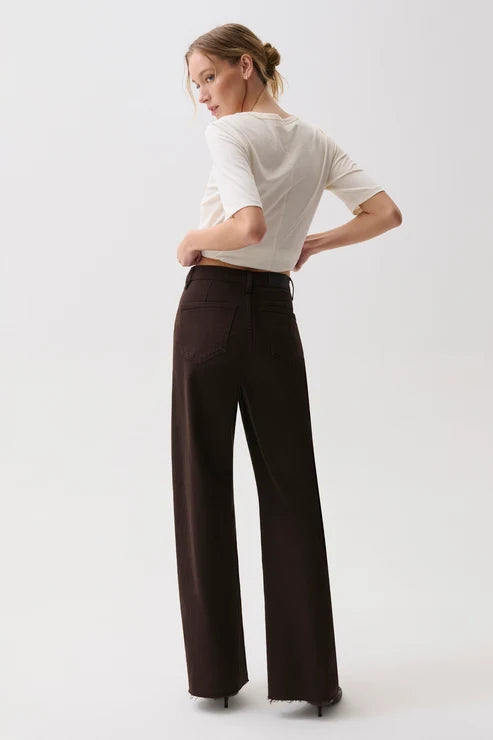 A woman wearing a white short-sleeve top and dark brown wide-leg trousers, photographed from the back against a plain light gray studio background.