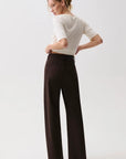 A woman wearing a white short-sleeve top and dark brown wide-leg trousers, photographed from the back against a plain light gray studio background.