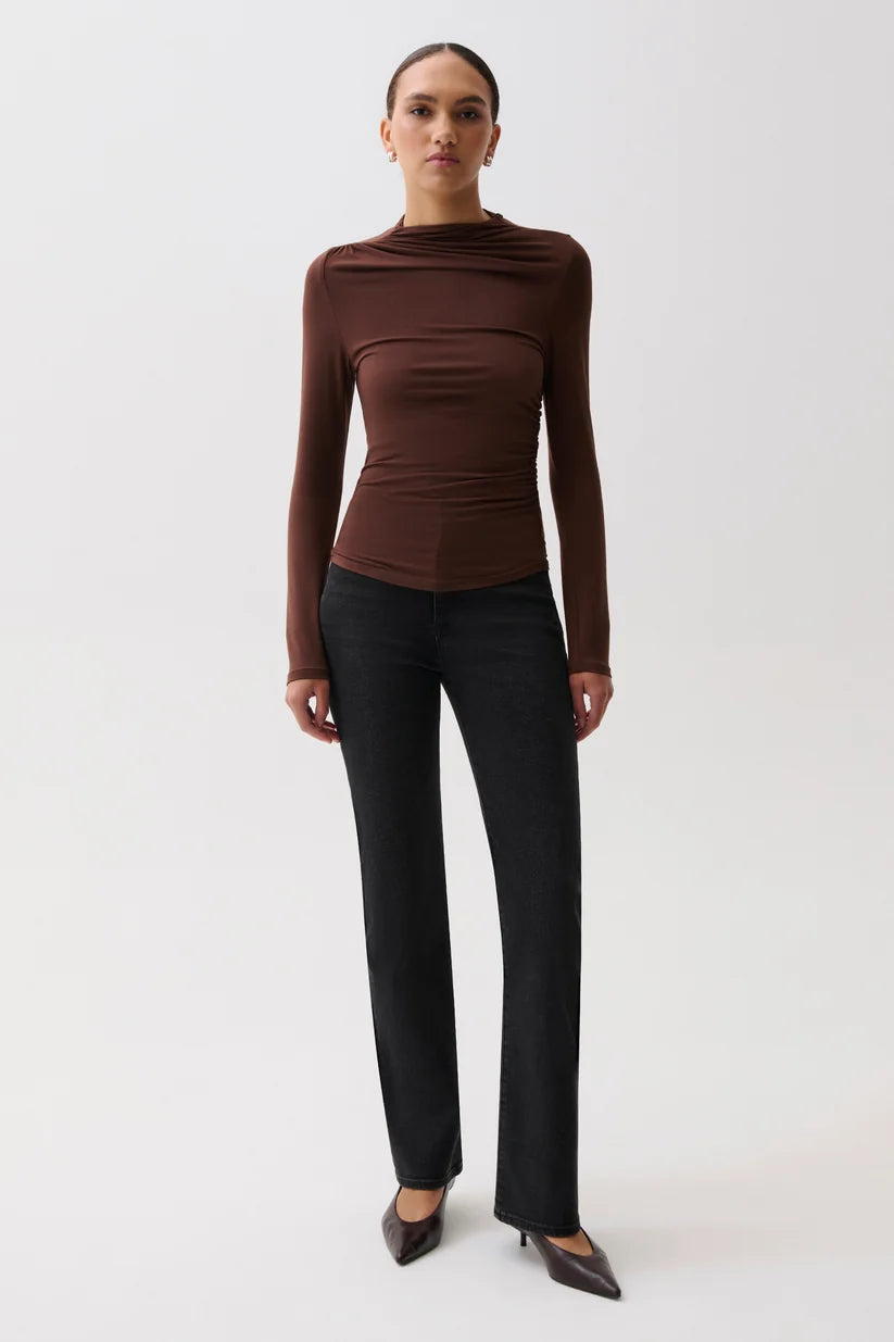 A model stands against a light gray studio background wearing a fitted, ruched long-sleeve top in dark brown with black trousers and dark pointed flats.