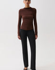 A model stands against a light gray studio background wearing a fitted, ruched long-sleeve top in dark brown with black trousers and dark pointed flats.
