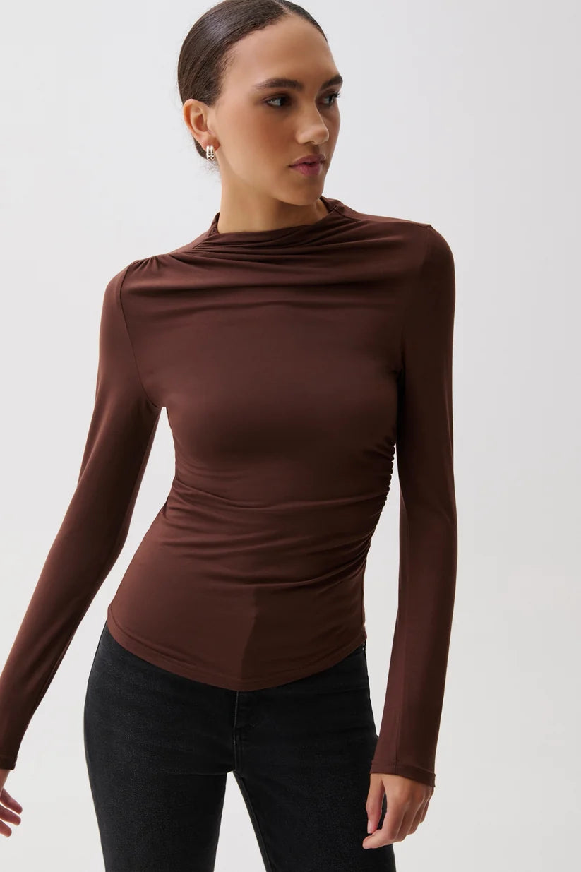 A woman wearing a fitted, long-sleeve brown top with a draped, ruched neckline and side details, paired with black pants, against a plain light gray background.