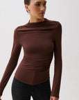 A woman wearing a fitted, long-sleeve brown top with a draped, ruched neckline and side details, paired with black pants, against a plain light gray background.