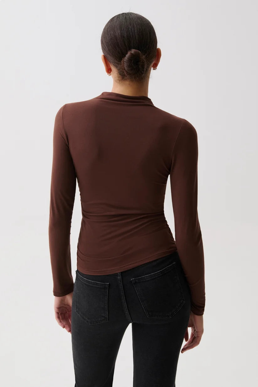 A woman viewed from the back wearing a fitted long-sleeve mock neck top in dark brown and black jeans, against a plain light gray studio background.