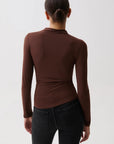 A woman viewed from the back wearing a fitted long-sleeve mock neck top in dark brown and black jeans, against a plain light gray studio background.