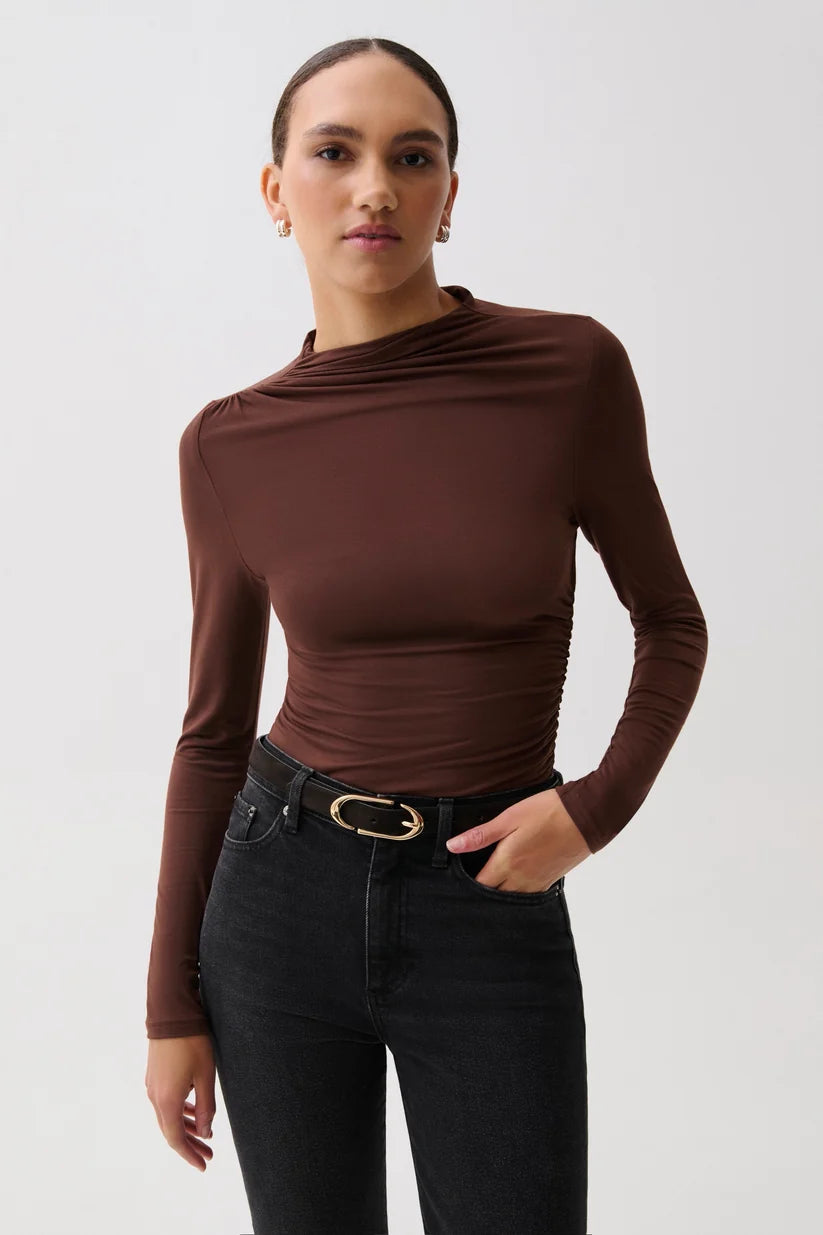 A woman modeling a dark brown long-sleeve top with a draped, asymmetrical neckline and gathered waist details, paired with black jeans and a black belt with a gold buckle, against a light gray studio background.