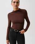 A woman modeling a dark brown long-sleeve top with a draped, asymmetrical neckline and gathered waist details, paired with black jeans and a black belt with a gold buckle, against a light gray studio background.