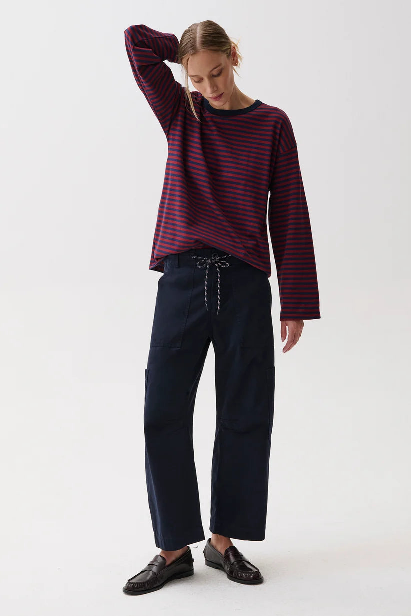 A woman modeling a dark red and navy blue horizontally striped long-sleeve top with dark navy wide-leg trousers and black leather loafers, standing against a plain white studio background.
