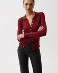 A woman modeling a fitted long-sleeve knit polo-style cardigan in deep burgundy with white buttons, paired with black pants, against a plain light gray studio background.