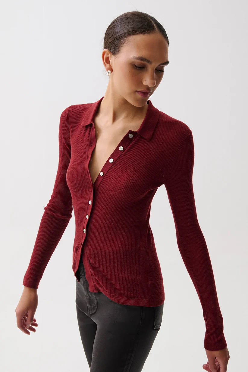 A woman wearing a fitted long-sleeve ribbed knit polo-style top in deep burgundy red, paired with dark gray pants, photographed against a clean light gray studio background.