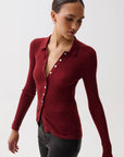 A woman wearing a fitted long-sleeve ribbed knit polo-style top in deep burgundy red, paired with dark gray pants, photographed against a clean light gray studio background.