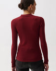 A woman shown from the back wearing a fitted long-sleeve ribbed knit top in deep burgundy red, paired with black pants, against a plain light gray studio background.