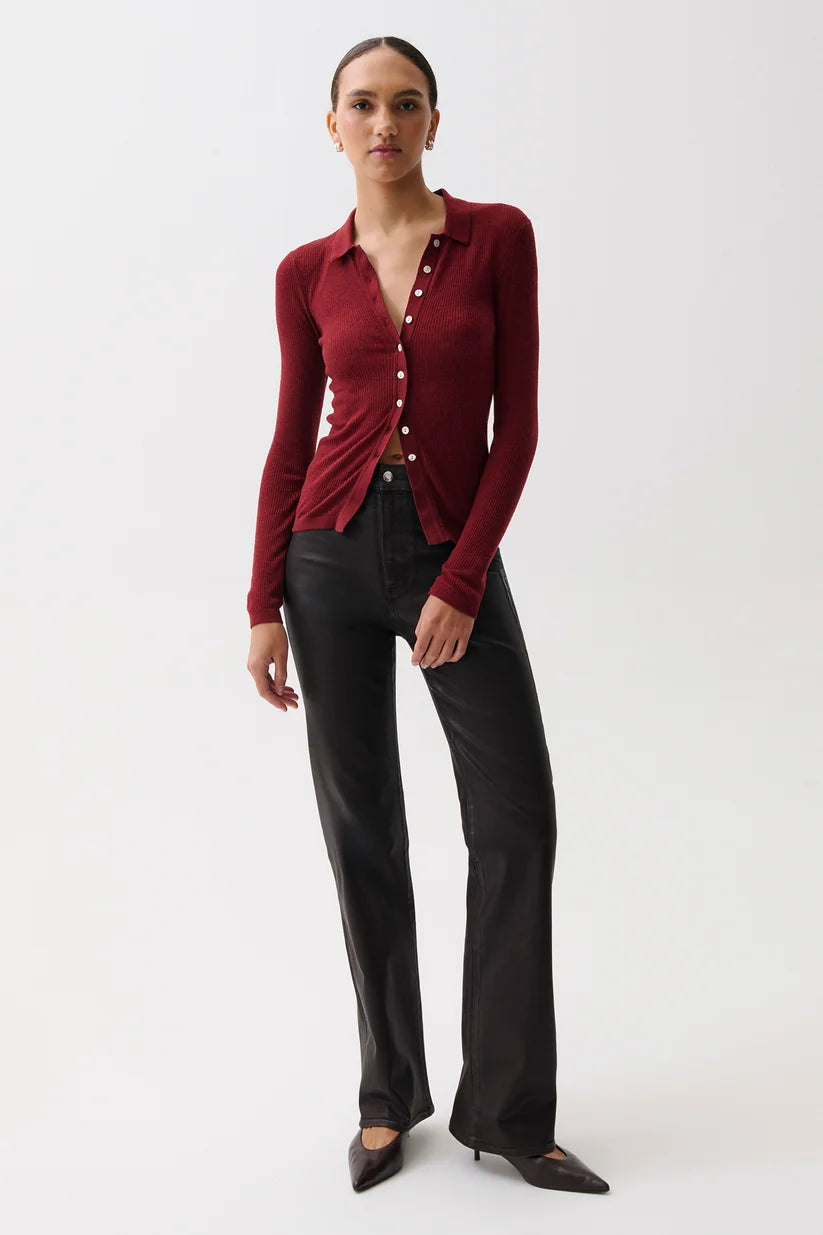 A model stands against a plain light gray studio background wearing a fitted long-sleeve burgundy button-front top with a twisted front detail, paired with black wide-leg pants and dark pointed flats.
