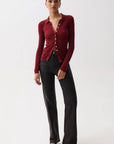A model stands against a plain light gray studio background wearing a fitted long-sleeve burgundy button-front top with a twisted front detail, paired with black wide-leg pants and dark pointed flats.