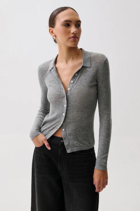 A woman in profile wearing a fitted gray ribbed knit polo cardigan with a buttoned collar over a black top and black pants, photographed against a light gray studio background.