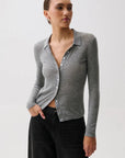 A woman in profile wearing a fitted gray ribbed knit polo cardigan with a buttoned collar over a black top and black pants, photographed against a light gray studio background.