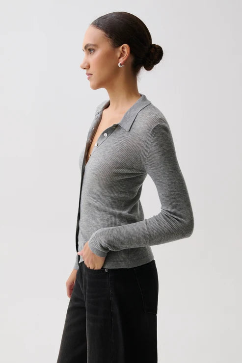 A woman in profile wearing a fitted gray ribbed knit polo cardigan with a buttoned collar over a black top and black pants, photographed against a light gray studio background.