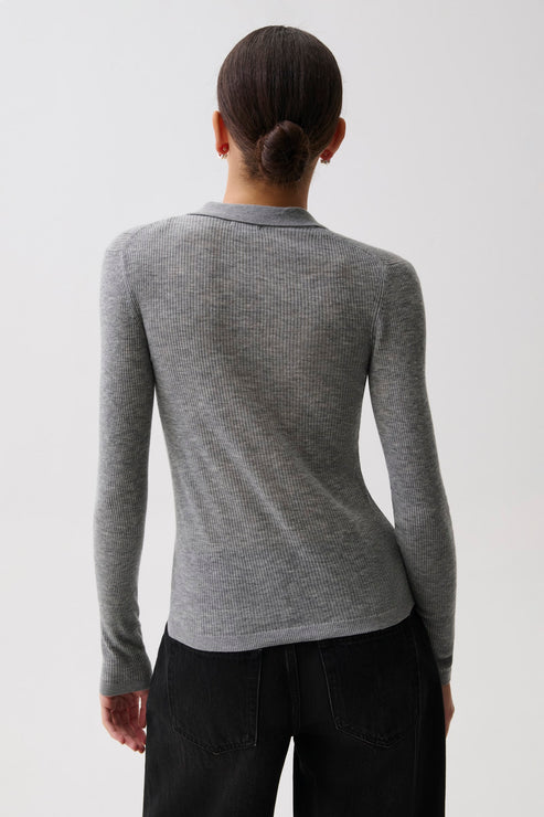 A woman in profile wearing a fitted gray ribbed knit polo cardigan with a buttoned collar over a black top and black pants, photographed against a light gray studio background, back