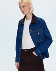 Woman wearing a blue denim jacket over a white shirt and black skirt on a light blue background, front/side view