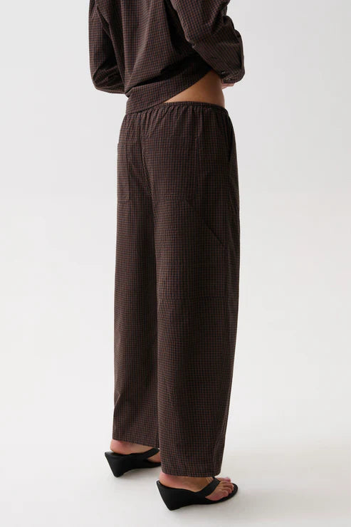 A person shown from the back wearing loose, wide-leg brown pants with a subtle small-scale check pattern, paired with black wedge sandals against a plain light gray studio background.