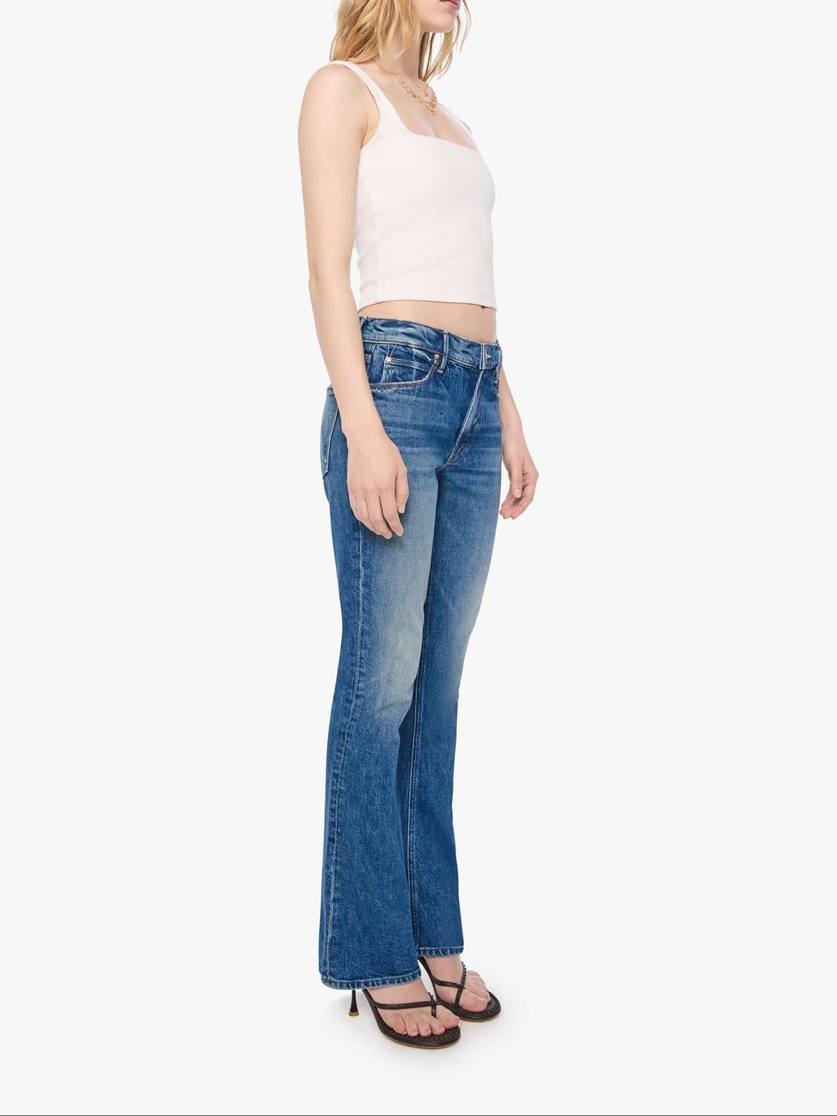 A woman modeling a pair of medium-wash blue flare jeans with a white ribbed crop top and sunglasses, standing against a plain white background.