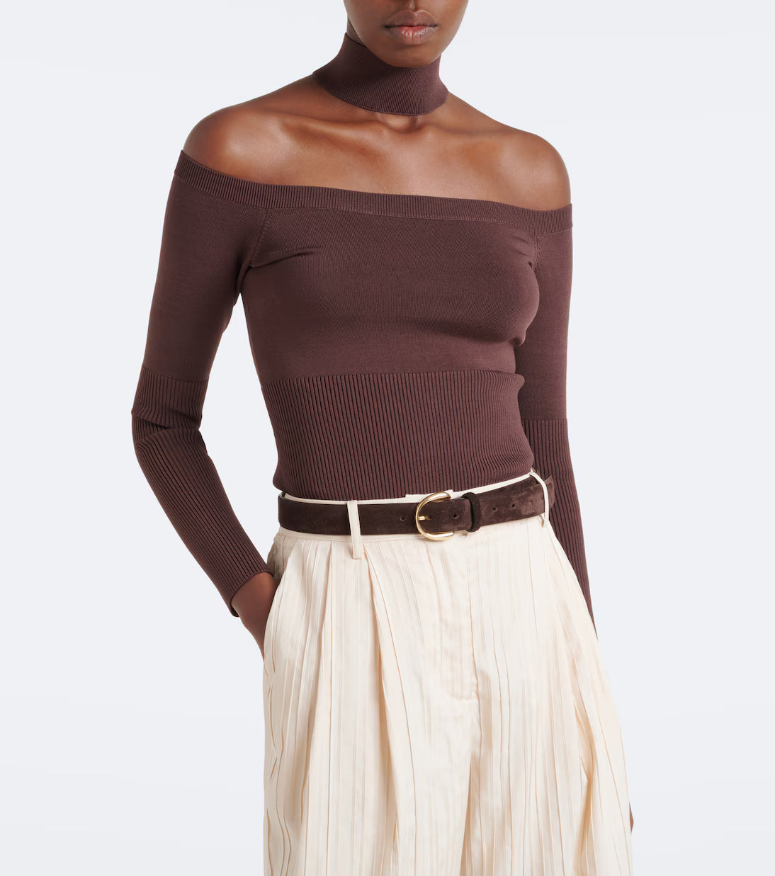 A model wearing a dark brown ribbed off-the-shoulder long-sleeve top paired with cream pleated trousers and a brown belt, shot against a plain white studio background.