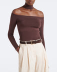 A model wearing a dark brown ribbed off-the-shoulder long-sleeve top paired with cream pleated trousers and a brown belt, shot against a plain white studio background.