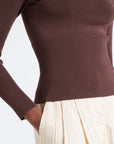 A close-up of a person wearing a fitted long-sleeve ribbed knit top in dark brown, paired with a cream pleated skirt, against a plain light background.