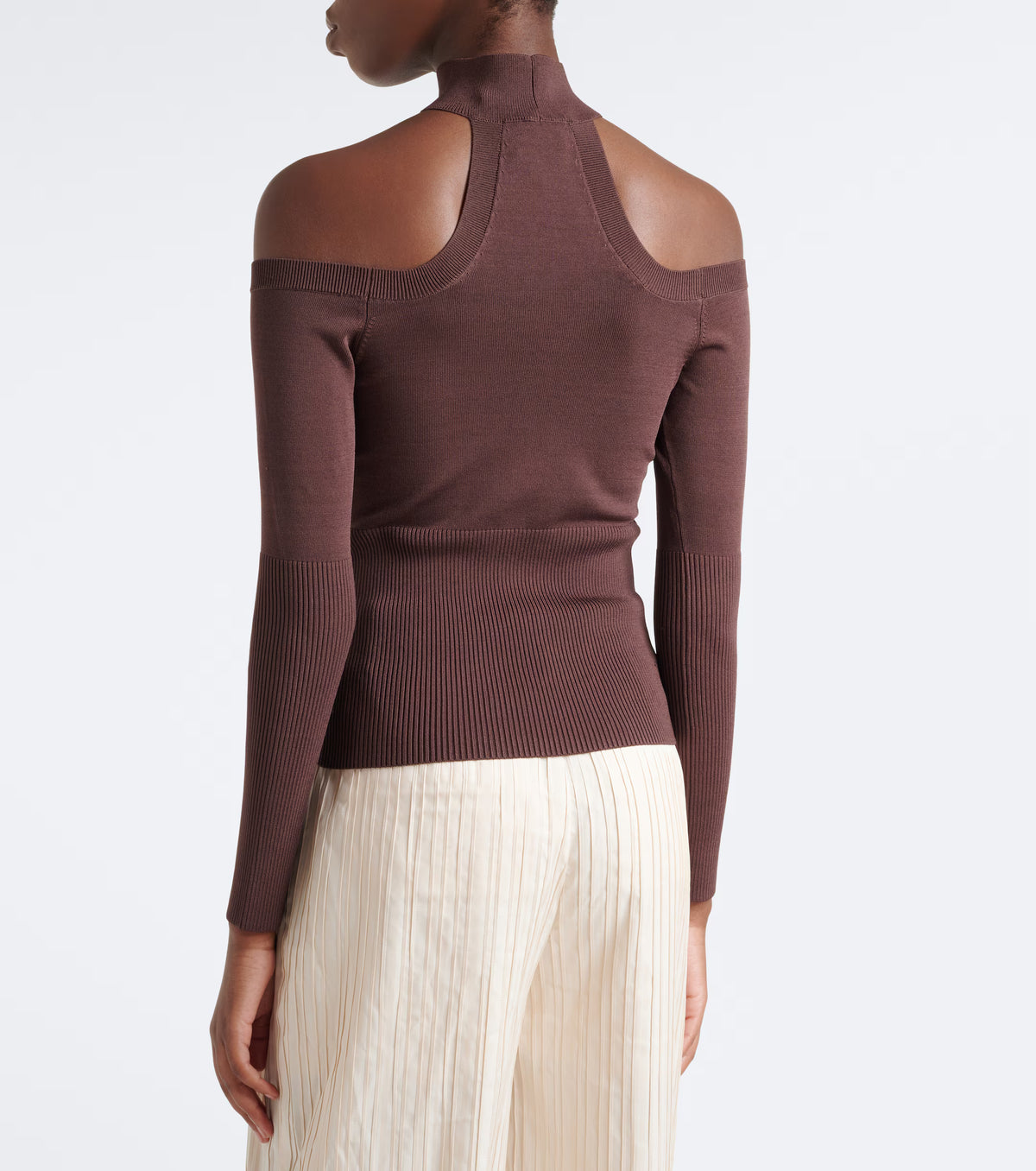A model shown from the back wearing a ribbed long-sleeve mock neck top with shoulder cutouts in a dark brown color, paired with cream pleated trousers, against a plain white background.