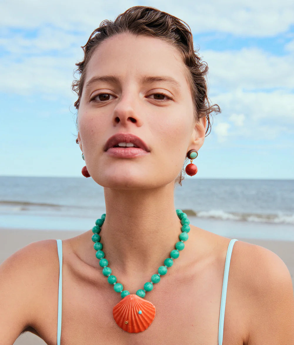 Woman wearing a turquoise necklace with a large orange pendant on a beach.
