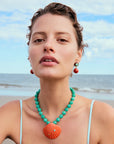 Woman wearing a turquoise necklace with a large orange pendant on a beach.