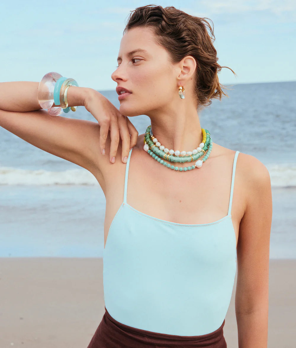 Woman on a beach wearing a light blue top and multiple necklaces.