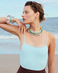 Woman on a beach wearing a light blue top and multiple necklaces.